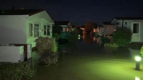 A collection of mobile homes at night surrounded by flood water. The scene is lit by green lights dotted around the site. 
