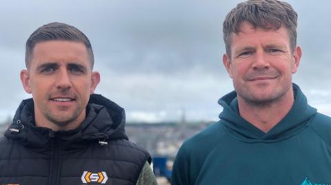 Darryl McDermott is wearing a black gilet with hood with a white and yellow coloured logo. He has short brown hair and stubble. 
Darryl McDermott, who is standing beside Dar