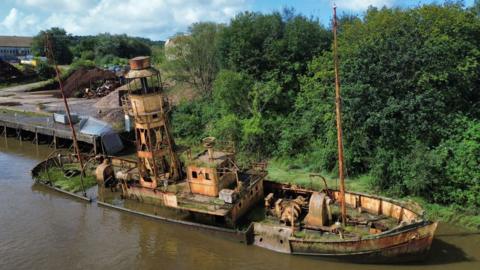A rusty ship with a light house attached to it. It is slightly leaning, with greenery behind it and muddy water in front.