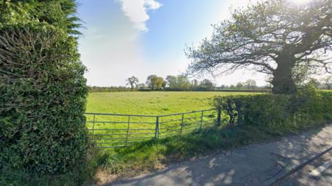 A Google street view image from a road showing a partially collapsed farm gate at the entrance to a large field surrounded by hedges and several trees.