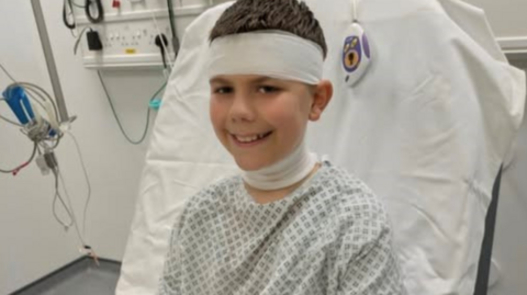 Olly is sitting up in bed in hospital wearing a patient's gown along with bandages on three right fingers, his neck and head. He is smiling