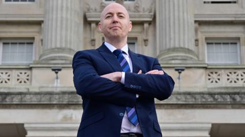 Jon Burrows looking at the camera with his arms folded, he is wearing a navy suit and blue tie.