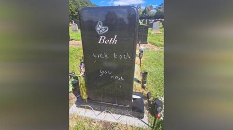 A photo of the grave which has the name 'Beth' engraved under a butterfly. The graffiti scrawled on the stone says 'tick tock you next'.