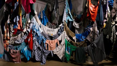 Shirts of different colours, designs and sizes are hung indoors around lines that drop down close to the floor where some of the shirts are touching the floor. There are hundreds of shirts on display.