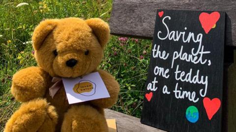 A teddy bear next to a sign that reads "Saving the planet, one teddy at a time"