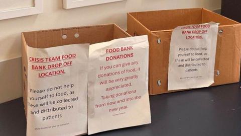Close up of two cardboard boxes with three pieces of white A4 paper taped to the front the papers read Crisis tram food bank drop off location and Food Bank Donations