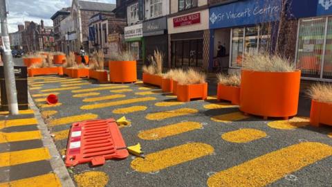 A road decorated with orange blobs painted on the tarmac. A bright orange barrier lies discarded on the carriageway. There are large orange planters along one side of the road, filled with dead grasses. There are shops along one side of the street. 