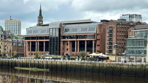 Newcastle Crown Court reflected in the River Tyne running in front of it. It is an imposing building made from smooth red stone with massive black windows and tall columns along its frontage.