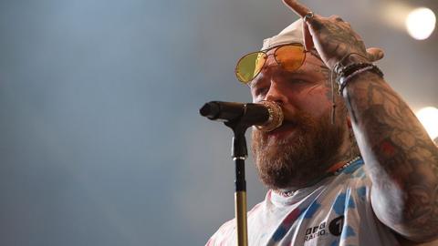 Teddy Swims wearing a cream cap and sunglasses resting on his forehead wearing a BBC Radio1 white t-shirt with blue and pink pattern performing at Radio 1's Big Weekend in July. 