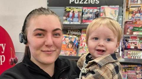 Molly Haggath is holding young Blake in her arms and smiling at the camera. They are in front of a magazine rack in the store, lined with colourful publications. Haggath is wearing a black uniform and a headpiece. She has brown hair which is pulled into a ponytail. Blake, who has short blonde hair and is wearing a brown and black jacket, has a joyful expression.