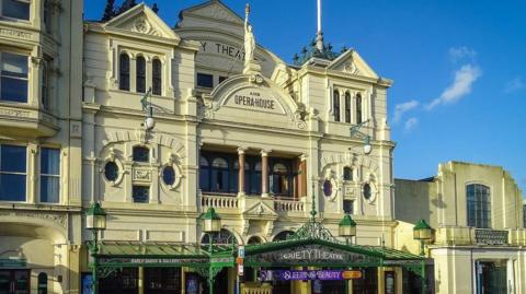 An ornate, Victorian-era theatre with a cream frontage. "Gaiety Theatre and opera house" is written in letters on the third floor, along with a greek statue of a woman holding a touch aloft. On the ground level there is a steel and glass overhanging shelter above the entrance.