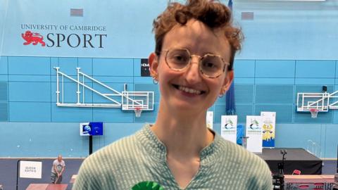 Kathryn Fisher standing on a balcony in a sports hall with desks laid out for an election count behind them. They have short hair, curling on the top and are wearing round glasses, a blue and white striped shirt and a green Green party rosette and are smiling broadly.