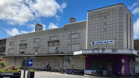 The derelict building in Bridgwater.  It has a largely grey exterior with boarded up windows and two chimneys on the top. There is a roundabout to the left of the image.