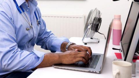 Doctor in a blue shirt with a stethescope round his neck looking at a laptop at a desk with a monitor. You can only see the body and not the head