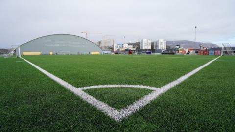 A low view of an artificial pitch, taken from one corner, looking towards high rise flats and a mountain in the distance