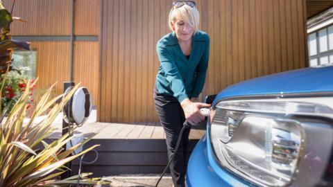 Woman charges blue electric car in her garden with decking and wooden walls seen behind her