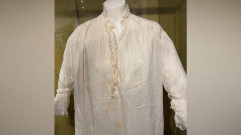 An old nightdress. It is white but has areas of yellowing. It is in a glass display case.