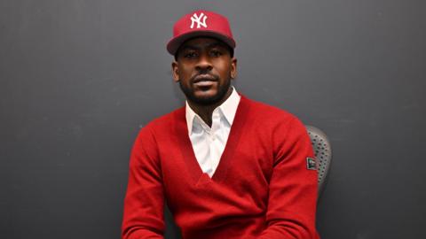 A UK grime artist wearing a red jumper with a white shirt underneath. He is wearing a red cap and looking directly at the camera.