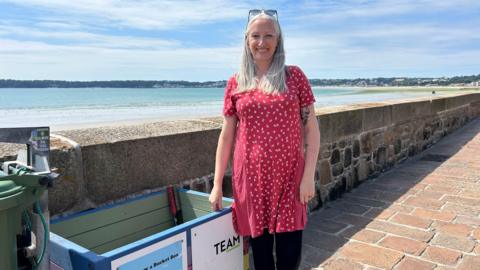 Louise Carson is wearing a red dress and stood on the promenade by St Aubin's Bay.