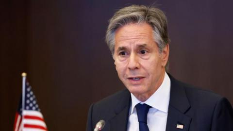 Close up shot of Antony Blinken speaking into a microphone. He is wearing a dark blue jacket, white shirt and dark blue tie. In the back