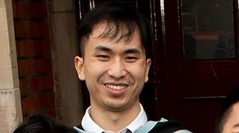 Zhenhao Zou is seen from the shoulders up in a cropped group picture. He is smiling wearing a graduation gown. 