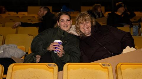 Two women sit in yellow chairs in a stadium stand, wearing dark waterproof jackets. They are huddled together and smiling, and the one on the left is holding a coffee cup. The woman on the left has dark hair tied back and the one on the right is blonde. It is dark but the stand is lit.