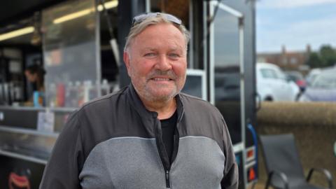 Glenn Dickerson is wearing a black and grey zip-up top. He's standing in front of a black food van.