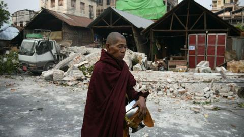 A monk in red robes walks past damaged houses and debris