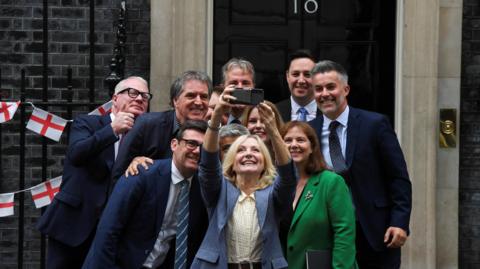 The regional mayors in England huddle together for a selfie in front of Number 10 Downing Street. A smiling Tracy Brabin holds her phone up to take the picture, with Andy Burnham to her right. There is some St George's cross bunting on the railings behind.