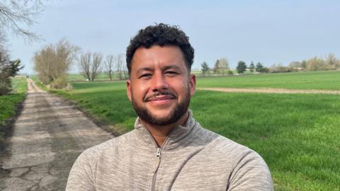 Ben Brooke has short, neat dark hair, a bit curly on top, he has a matching beard and moustache and is wearing a grey cotton sports top zipped up to his neck. Behind him are grass fields with a rough track wide enough for one vehicle running through it.