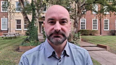 A bald man with a dark greying beard and wearing a blue-grey collared shirt. In the background there is a row of buildings and trees.