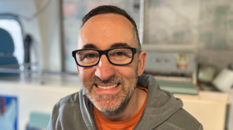 A man in dark rimmed spectacles, short dark hair and a grey speckled beard and moustache is looking at the camera and smiling. 