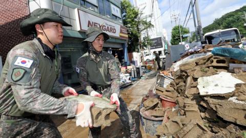 Two South Korean soldiers in camouflage military uniforms remove a large piece of rock from a pile of fallen debris on an urban street.