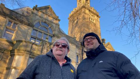 Two men, one with silver hair and wearing dark sunglass, the other in clear glasses and woolly hat standing outside the Yorkshire stone hall 