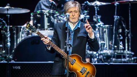Paul McCartney on stage wearing a blue jacket, blue shirt and with a guitar. He is pointing one finger in the hair.