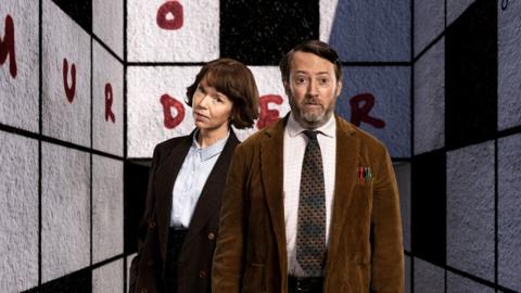 David Mitchell and Anna Maxwell Martin looking at the camera in a black and white square tiled room with the word murder written on the wall