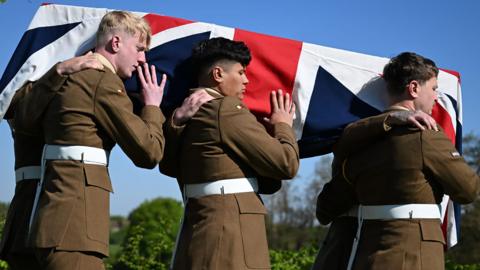 A side view of the Union Jack draped coffin holding the remains of Private Reginald Joseph Blake at the Commonwealth War Graves Commission's Loos British Cemetery Extension, on 22 April 2026. The coffin is held on the shoulders of six Royal Anglian Regiment soldiers, three of whom can be seen. Behind is blue sky and green trees. 