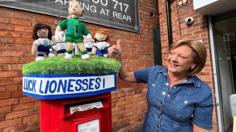 A woman wearing a denim top stands next to a postbox topper and gives a thumbs-up sign. She has a short bob hairstyle. The topper has a knitted figure of England goalie Hannah Hampton wearing a green kit in front of several other players in blue and white kits.