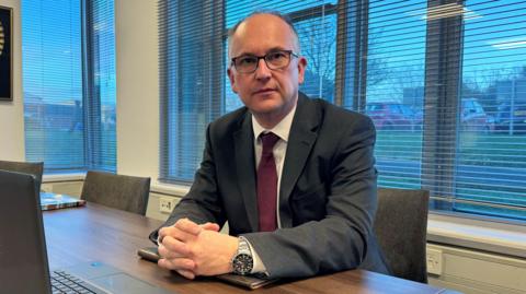 A man sat behind a desk, wearing a black suit, white shirt and red tie. He is in front of a laptop.