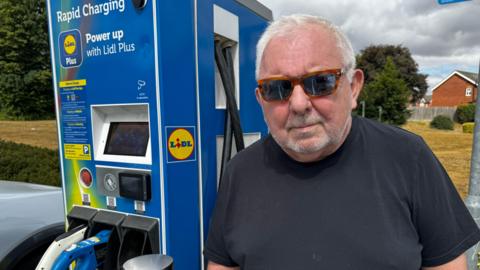 A man wearing sunglasses and a black T-shirt stands beside a blue Lidl Plus rapid electric vehicle charging unit at Lidl's Barton-upon-Humber store. Charging cables are connected to a vehicle parked alongside the charger. The unit displays instructions for using the charging service, while trees, housing and dark clouds are visible in the background.