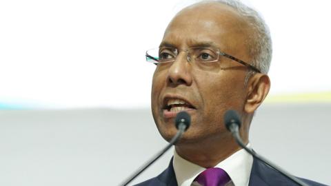 Lutfur Rahman at an official event, speaking into two small microphones. He wears a suit and a purple tie