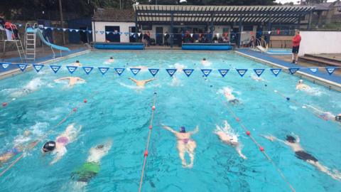 About a dozen people are swimming in the six-lane outdoor pool. They are wearing swimsuits and caps in varying colours. There is blue bunting above them and a lifeguard on either side of the pool. Ahead, more people are under a veranda and watching the swimmers.