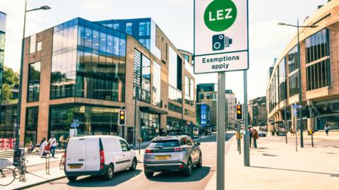 a sign that reads low emission zone exemptions apply on a road in edinburgh with two cars waiting at traffic lights nearby and pedestrians walking