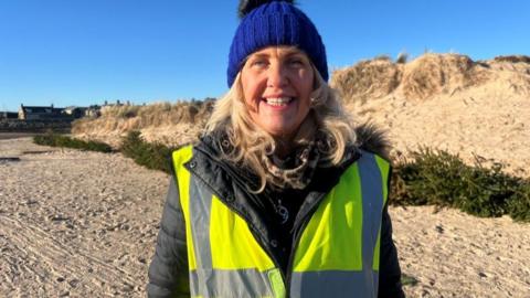 Carolle Ralph, woman with blonde hair smiling at camera, wearing hi-vis yellow jacket and warm blue hat, on a sandy beach with Christmas trees lying on the ground in front of dunes.