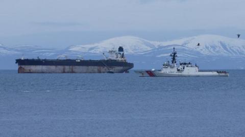 The tanker is a large ship with rusty marks on its hull. The upper part of its hull is painted blue and it has a white bridge and a single blue funnel. In the foreground is a white US Coast Guard vessel. There are snow-covered hills in the background.