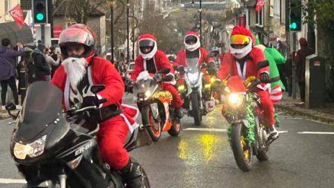 A group of bikers, four are dressed as Santa, one as the Grinch. The Tamar Bridge can be seen in the background and there are crowds lining the street.