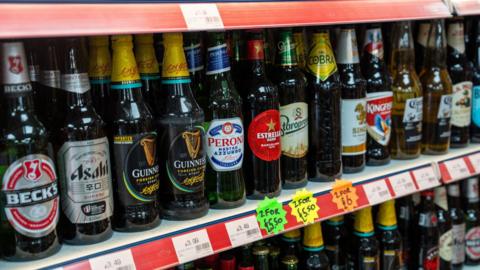 A shop shelf filled with rows of assorted bottled beers from various brands, each with price labels and multibuy offer signs displayed beneath them.