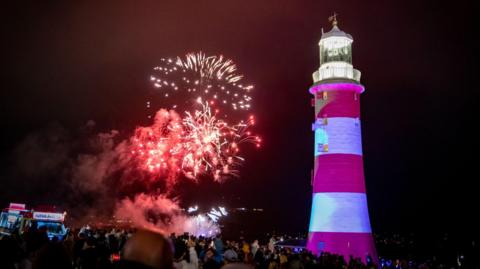 A number of fireworks explode behind Smeaton's Tower. The tower is red and white striped with a lantern at the top.