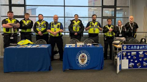Nine West Midlands police staff members line up behind blue tables and in front of a window. Two of the tables are covered in blue table clothes and a second behind a careers pinboard. Seven are wearing police uniform and yellow high-vis jackets with walkie talkies attached. Two pictured are women.