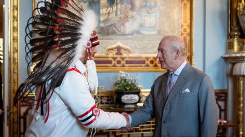 An image showing Grand Chief Joey Pete shaking hands with King Charles III in Buckingham Palace. Behind them is an ornate backdrop featuring gold furniture and a large painting, with light blue walls. Grand Chief Pete (on the left) is wearing a ceremonial indigenous headdress. King Charles is wearing a grey suit. 